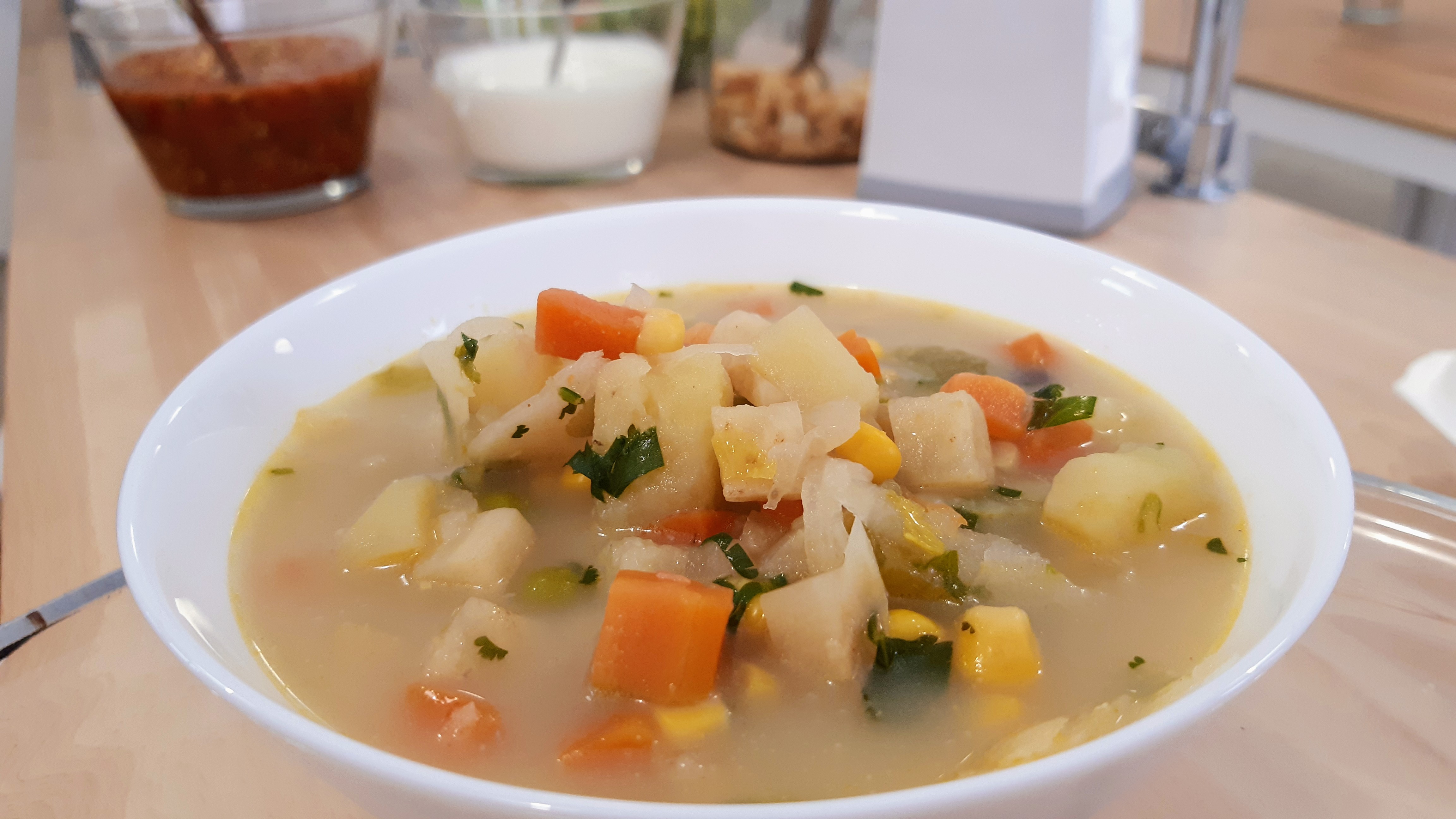 Porcja zupy jarzynowej w białej misce, z widocznymi kawałkami ziemniaków, marchwi, kukurydzy i zieleniny, serwowana z dodatkami w przezroczystych naczyniach w tle.