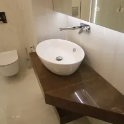 Nowoczesna łazienka z białą owalną umywalką nablatową, chromowaną armaturą, brązowym blatem z efektem marmuru, płytkami w odcieniach bieli ułożonymi w jodełkę oraz toaletą w tle.