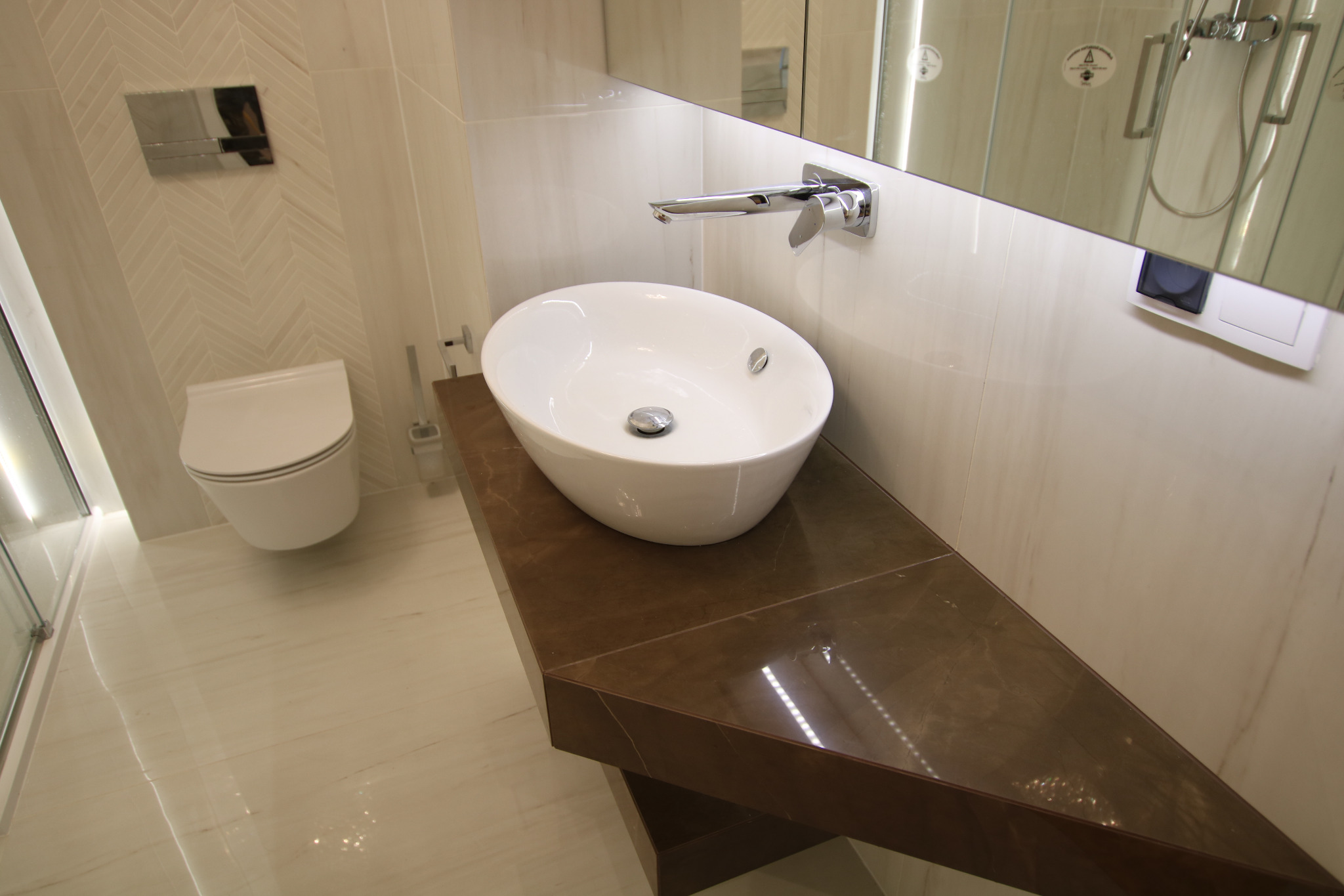 Nowoczesna łazienka z białą owalną umywalką nablatową, chromowaną armaturą, brązowym blatem z efektem marmuru, płytkami w odcieniach bieli ułożonymi w jodełkę oraz toaletą w tle.
