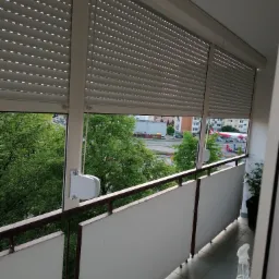 Szare rolety zewnętrzne na balkonie z widokiem na zieleń i stację benzynową; mechanizm zwijania rolet widoczny po prawej stronie.