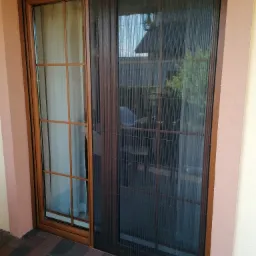 Brązowa moskitiera plisowana zamontowana na ramie okiennej z widoczną firanką w tle.