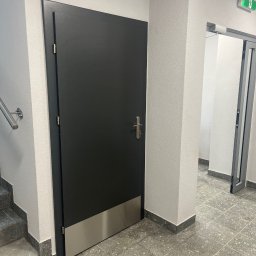 FPHU DOOR WIT Dawid Wituła - Ciemnoszare drzwi z metalowym panelem dolnym, zamontowane w klatce schodowej budynku, widoczny fragment schodów i drzwi ewakuacyjne w tle.