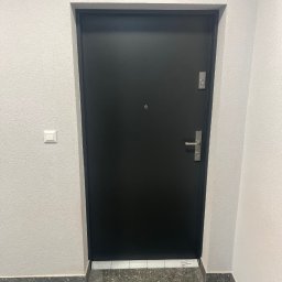 FPHU DOOR WIT Dawid Wituła - Czarna, gładka, solidna brama wejściowa z srebrną klamką i zamkiem, zamontowana w jasnym korytarzu z szarą podłogą i włącznikiem światła po lewej stronie.
