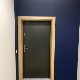 FPHU DOOR WIT Dawid Wituła - Ciemnoszare drzwi wejściowe z jasną ościeżnicą, zamontowane w korytarzu z białą i granatową ścianą. Na podłodze widoczne ślady po remoncie.