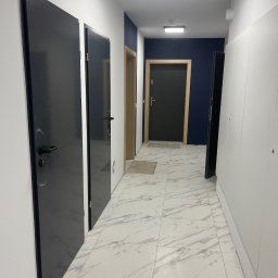 FPHU DOOR WIT Dawid Wituła - Wnętrze korytarza z trzema parami drzwi: ciemnoszare drzwi wejściowe z czarną klamką po lewej, jasne drzwi wewnętrzne w środku i ciemnoszare drzwi na końcu korytarza, białe ściany i marmurowa podłoga.