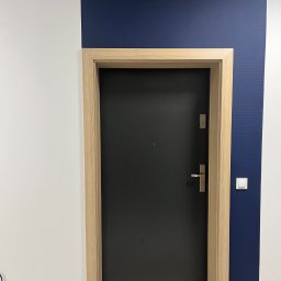 FPHU DOOR WIT Dawid Wituła - Czarna, nowoczesna brama wejściowa z jasną, drewnianą futryną na tle białej i granatowej ściany z włącznikiem światła.