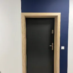 Czarna, nowoczesna brama wejściowa z jasną, drewnianą futryną na tle białej i granatowej ściany z włącznikiem światła.