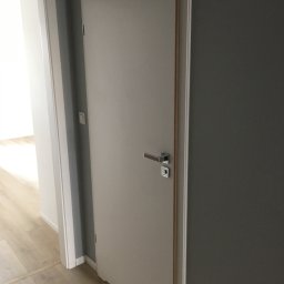 FPHU DOOR WIT Dawid Wituła - Zamknięte, gładkie, szare drzwi wewnętrzne z srebrną klamką, widoczne w korytarzu z drewnianą podłogą i otwartymi drzwiami w tle.