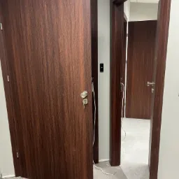 Brązowe drzwi wewnętrzne z widocznymi kablami podczas montażu, z kluczem w zamku, w tle widoczne kolejne drzwi i szara podłoga w trakcie wykończenia.