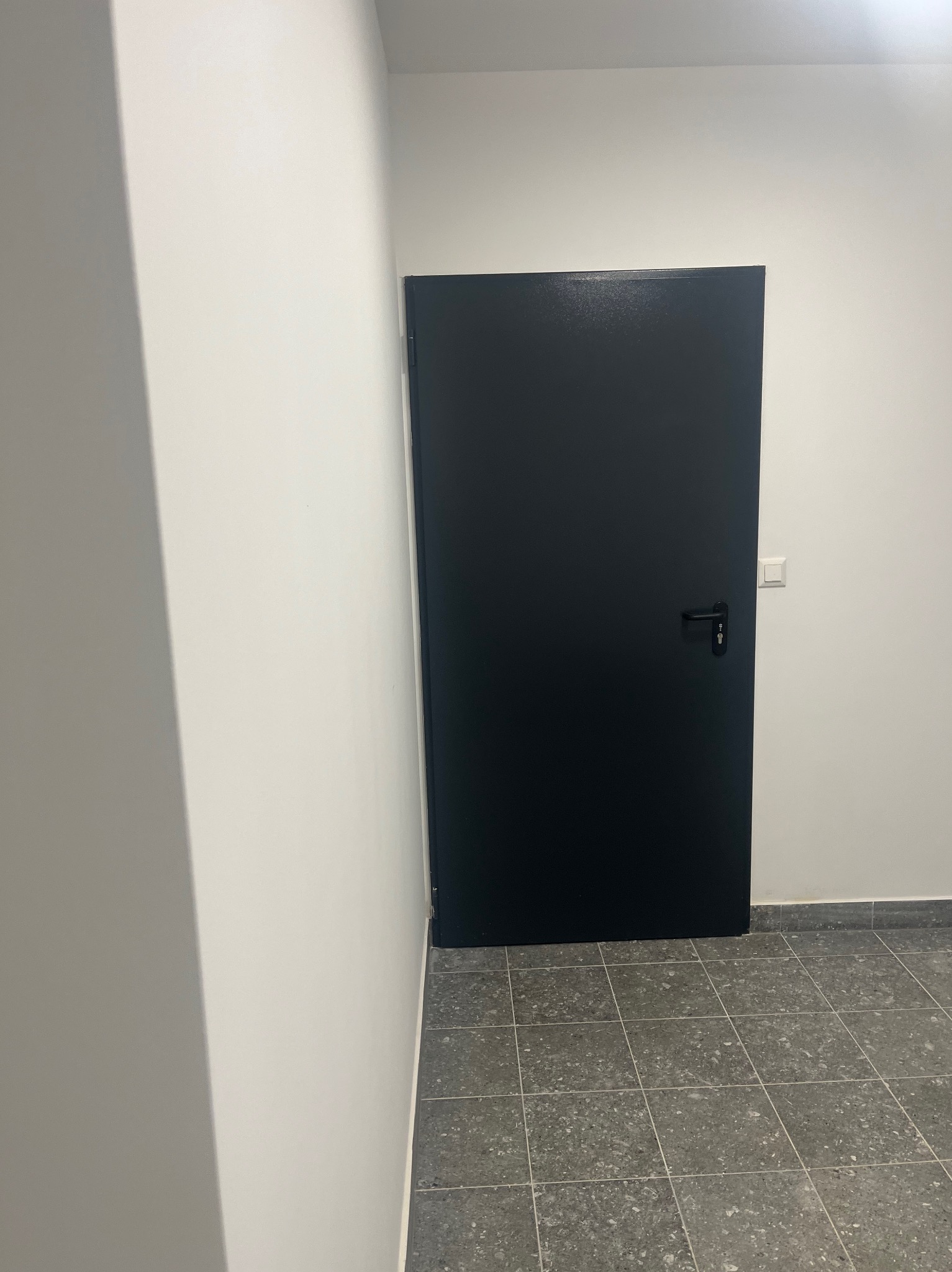 Zamknięte, czarne drzwi wewnętrzne z czarną klamką, umieszczone w białej ścianie, z widocznym włącznikiem światła i szarą podłogą z płytek.