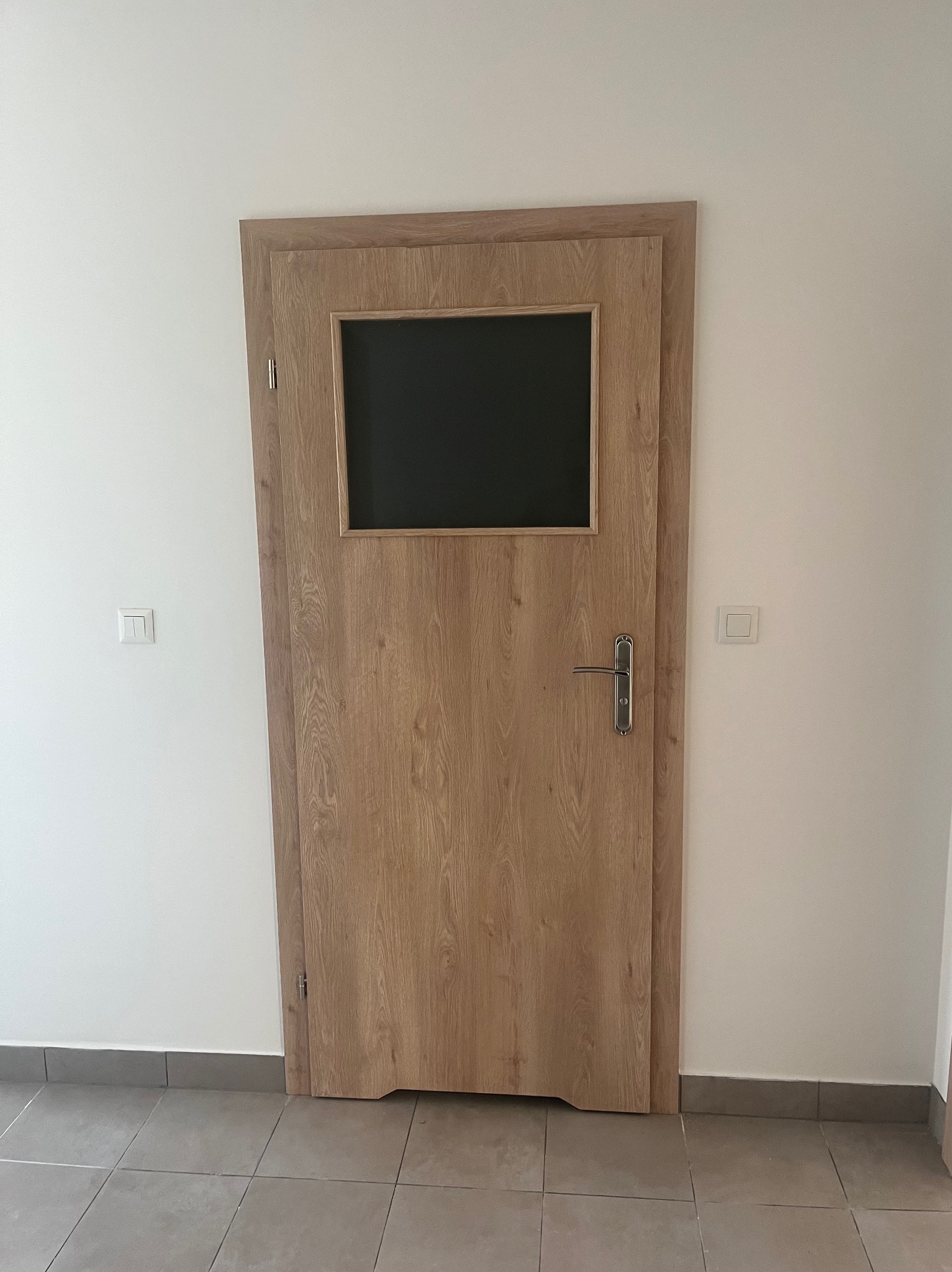 Zamknięte drzwi wewnętrzne z drewnianą ramą i prostokątnym przeszkleniem, srebrna klamka, widoczne płytki podłogowe i białe ściany z włącznikami.