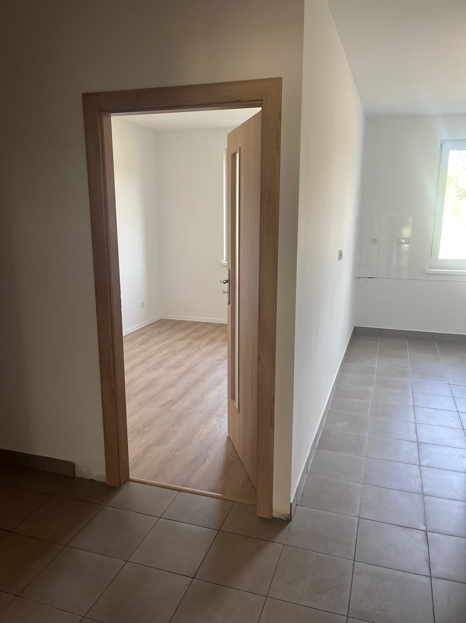 Otwarta, drewniana ościeżnica z częściowo przeszklonymi skrzydłami drzwiowymi, widok na puste, jasne pomieszczenie z panelami podłogowymi, szare płytki podłogowe w przyległym pokoju.