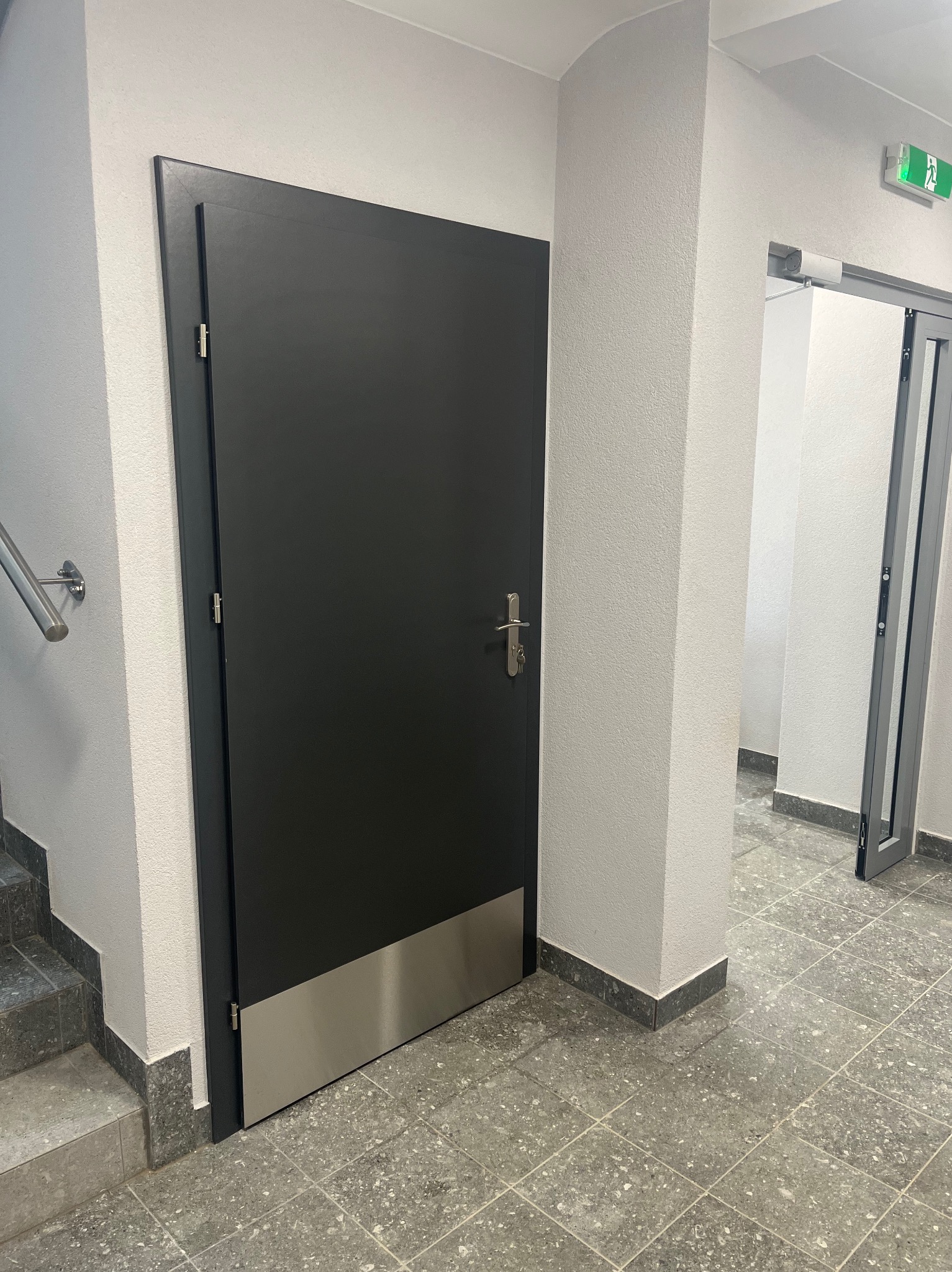 Ciemnoszare drzwi z metalowym panelem dolnym, zamontowane w klatce schodowej budynku, widoczny fragment schodów i drzwi ewakuacyjne w tle.