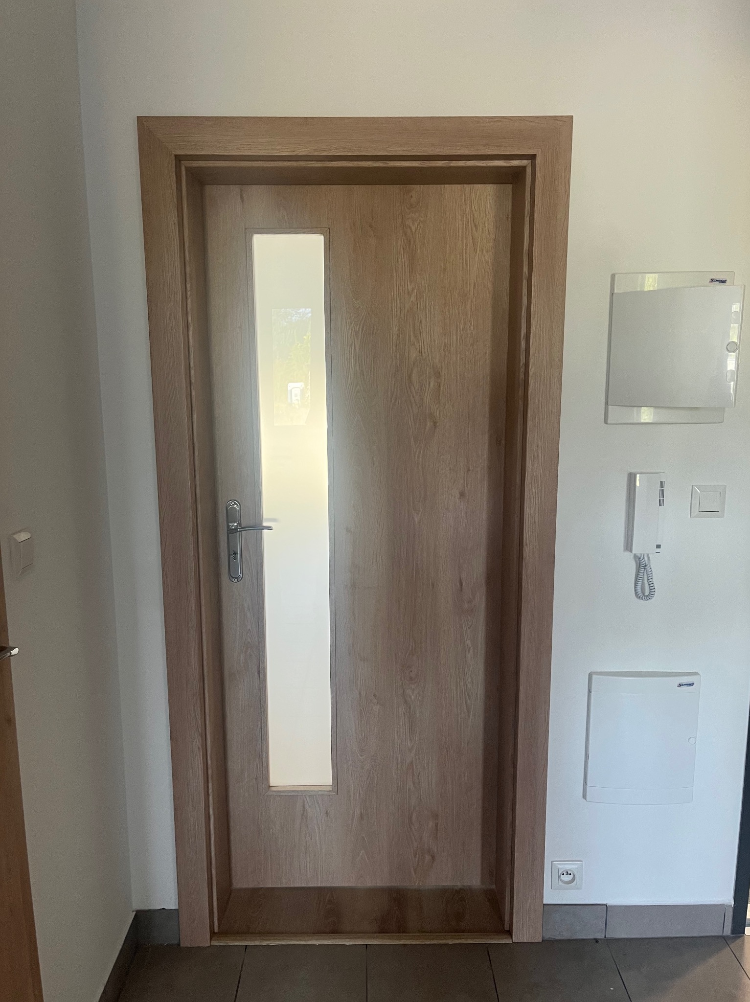 Drewniane drzwi wewnętrzne z prostokątnym przeszkleniem, srebrna klamka, widoczne elementy instalacji elektrycznej na ścianie.