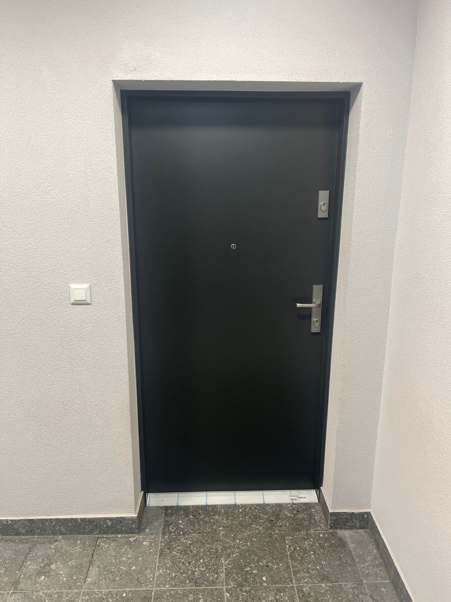 Czarna, gładka, solidna brama wejściowa z srebrną klamką i zamkiem, zamontowana w jasnym korytarzu z szarą podłogą i włącznikiem światła po lewej stronie.