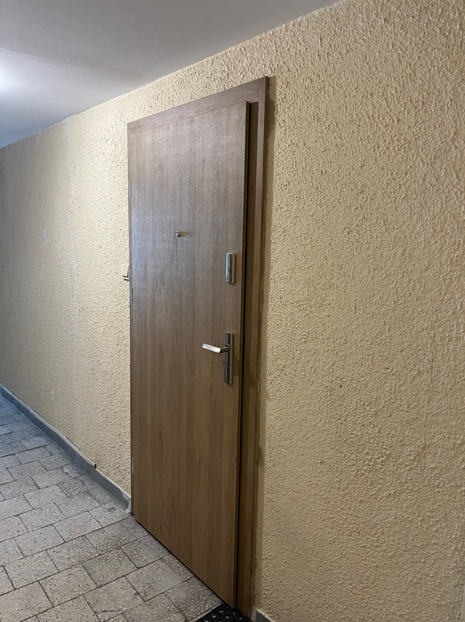 Drewniane drzwi wejściowe z wizjerem i srebrną klamką, zamontowane w korytarzu o beżowych ścianach i szarej podłodze z płytek.