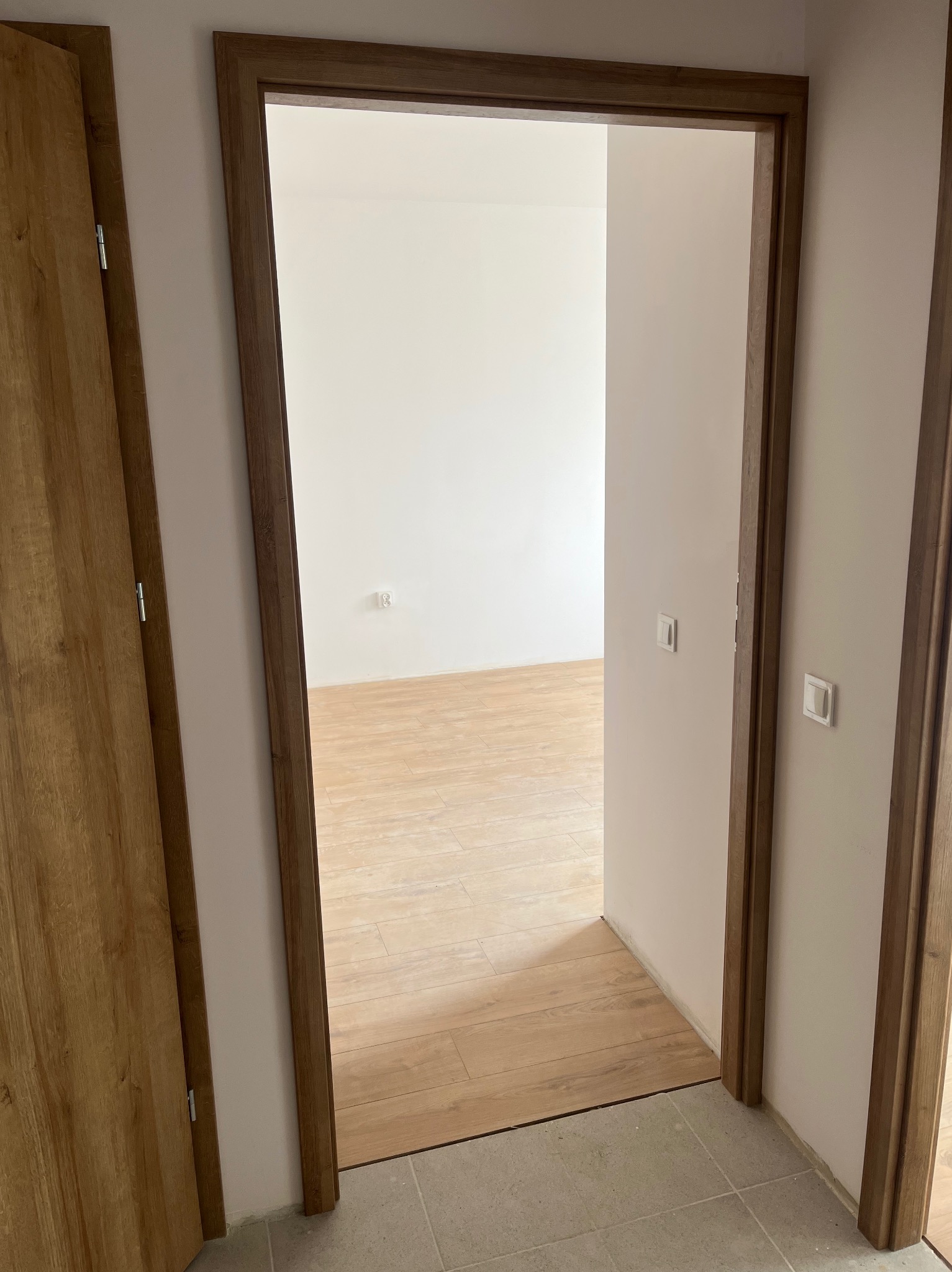 Otwarty widok na wnętrze mieszkania przez drewnianą futrynę i częściowo otwarte drzwi o podobnym wykończeniu, ukazujący jasne ściany i podłogę wyłożoną panelami.