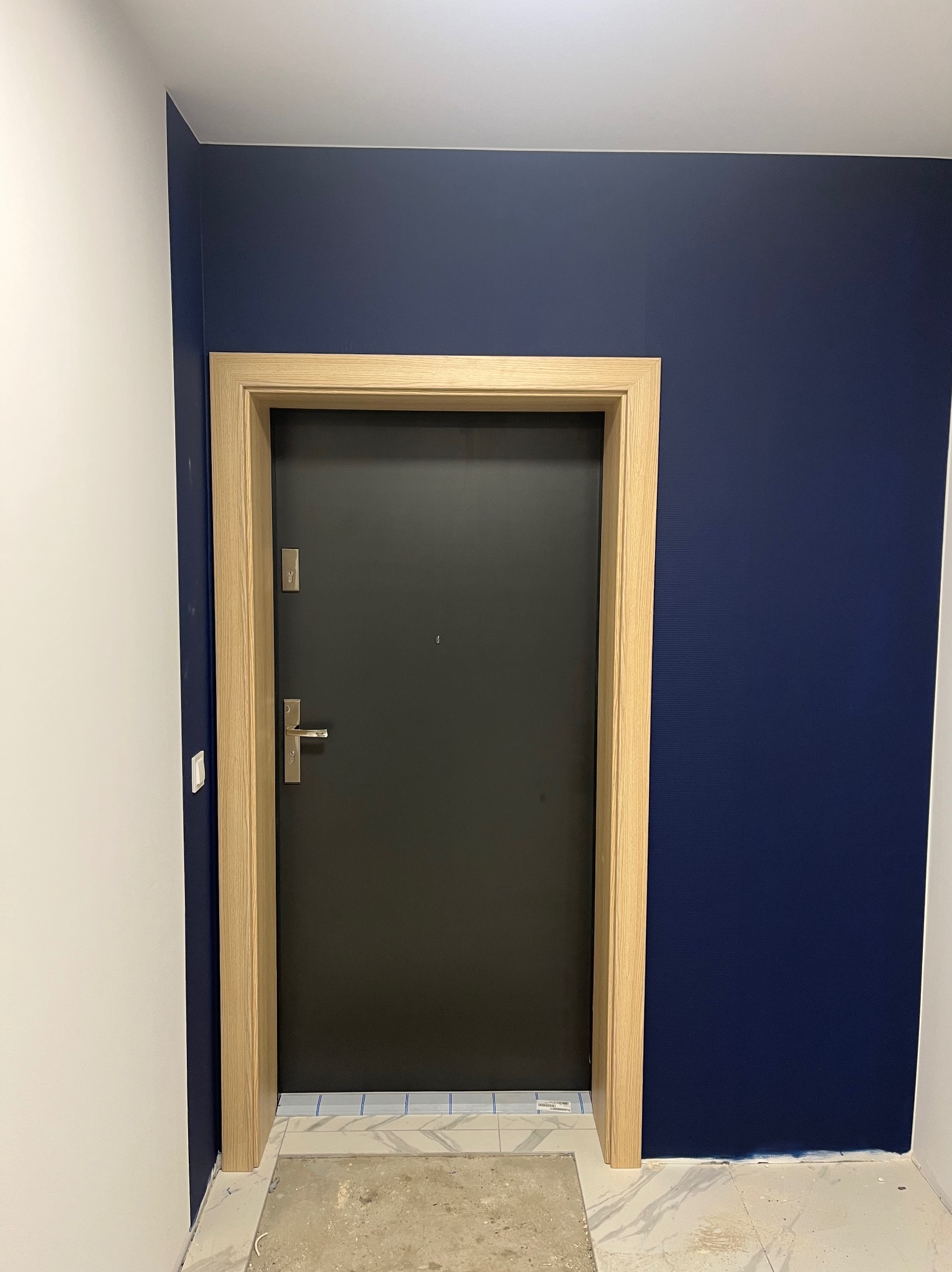 Ciemnoszare drzwi wejściowe z jasną ościeżnicą, zamontowane w korytarzu z białą i granatową ścianą. Na podłodze widoczne ślady po remoncie.