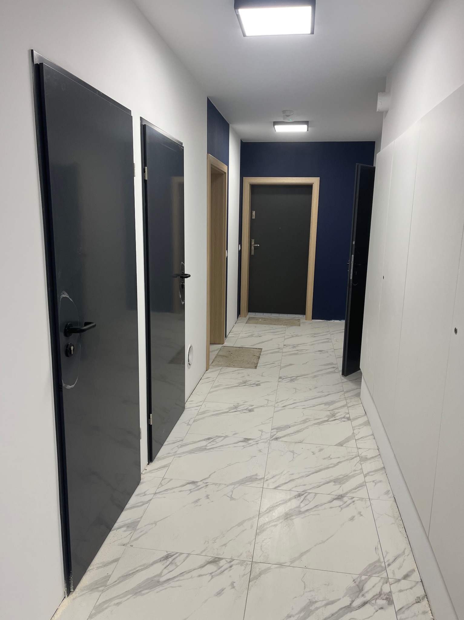 Wnętrze korytarza z trzema parami drzwi: ciemnoszare drzwi wejściowe z czarną klamką po lewej, jasne drzwi wewnętrzne w środku i ciemnoszare drzwi na końcu korytarza, białe ściany i marmurowa podłoga.