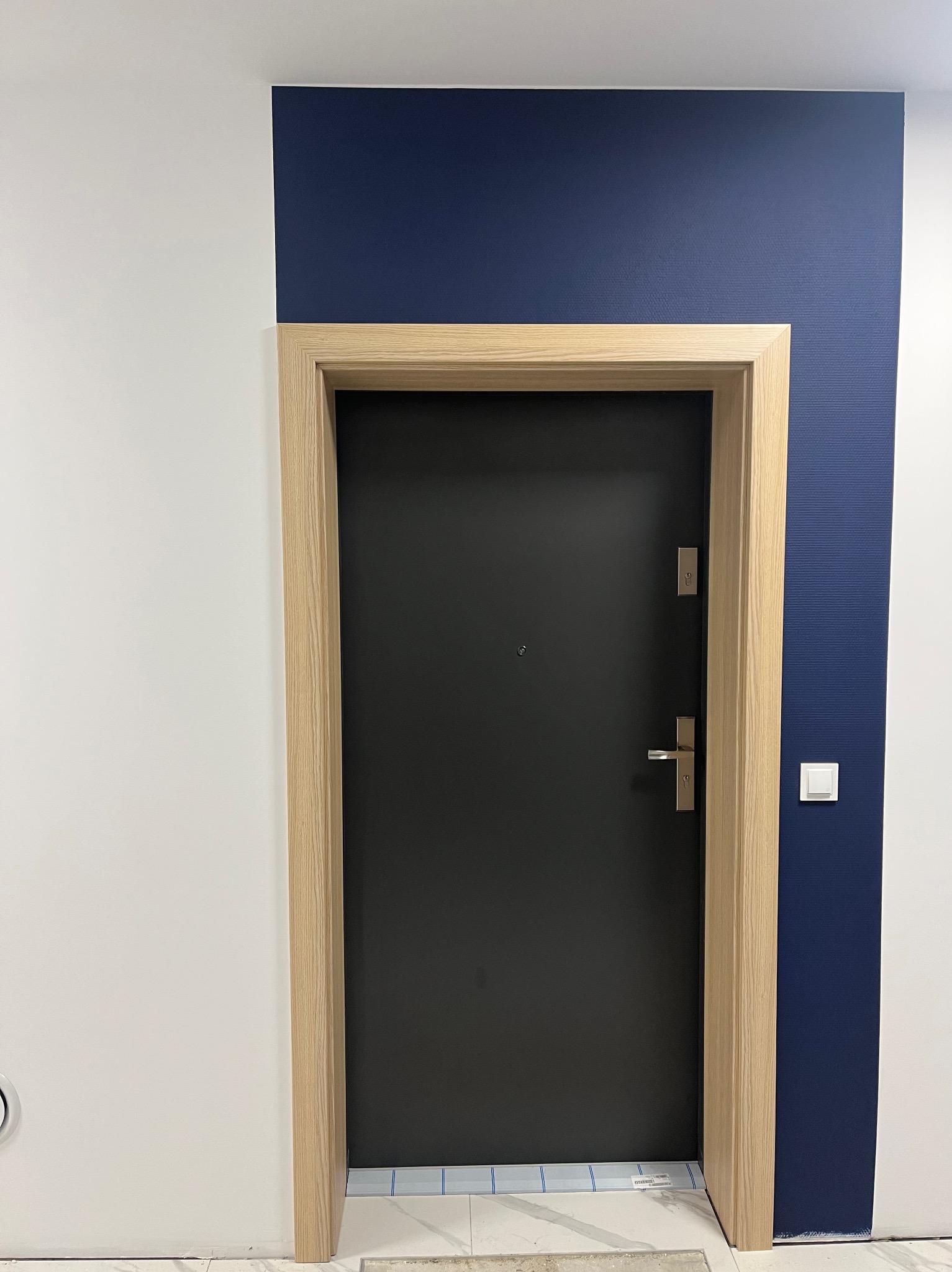 Czarna, nowoczesna brama wejściowa z jasną, drewnianą futryną na tle białej i granatowej ściany z włącznikiem światła.