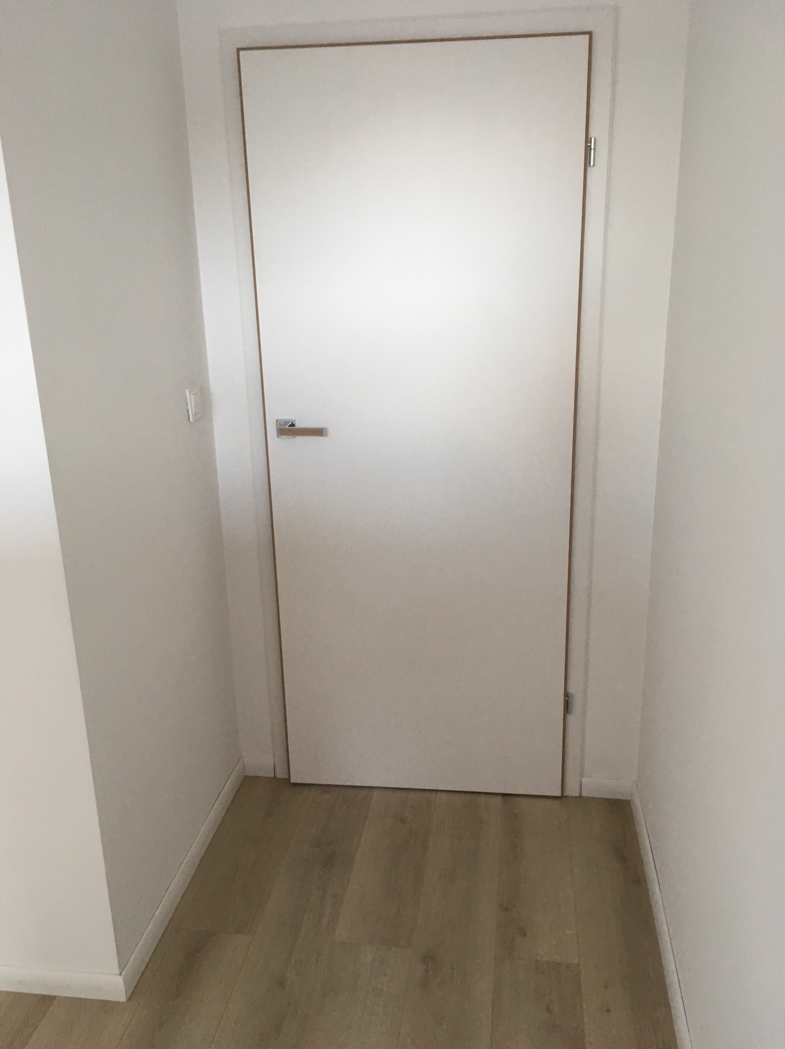 Białe drzwi wewnętrzne z klamką i ościeżnicą w jasnym korytarzu z panelami podłogowymi.