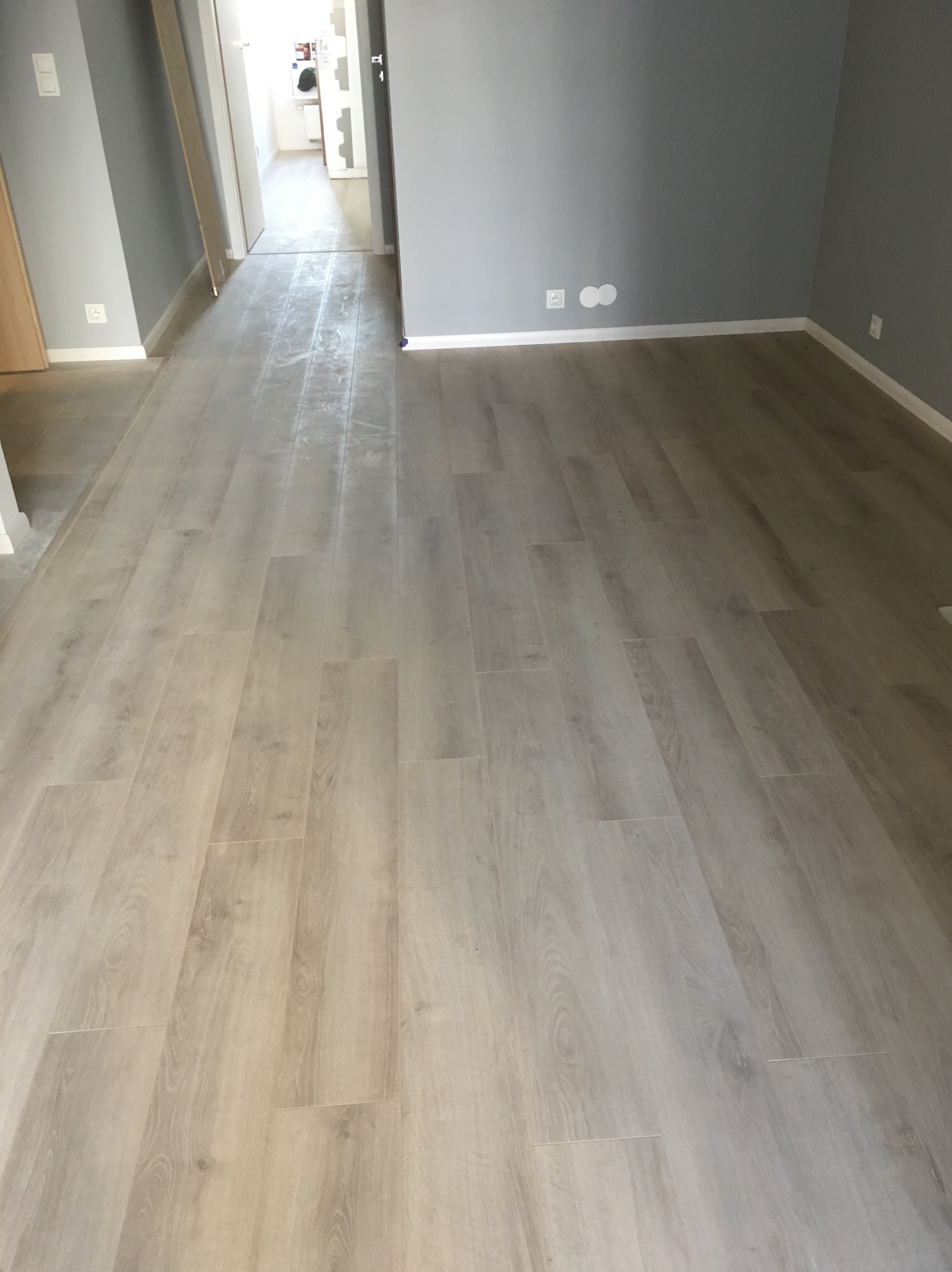 Jasna, drewnopodobna podłoga panelowa ułożona w pustym, szarym pokoju z widocznymi gniazdkami elektrycznymi przy listwach przypodłogowych. W tle widoczne otwarte drzwi do kolejnego pomieszczenia.