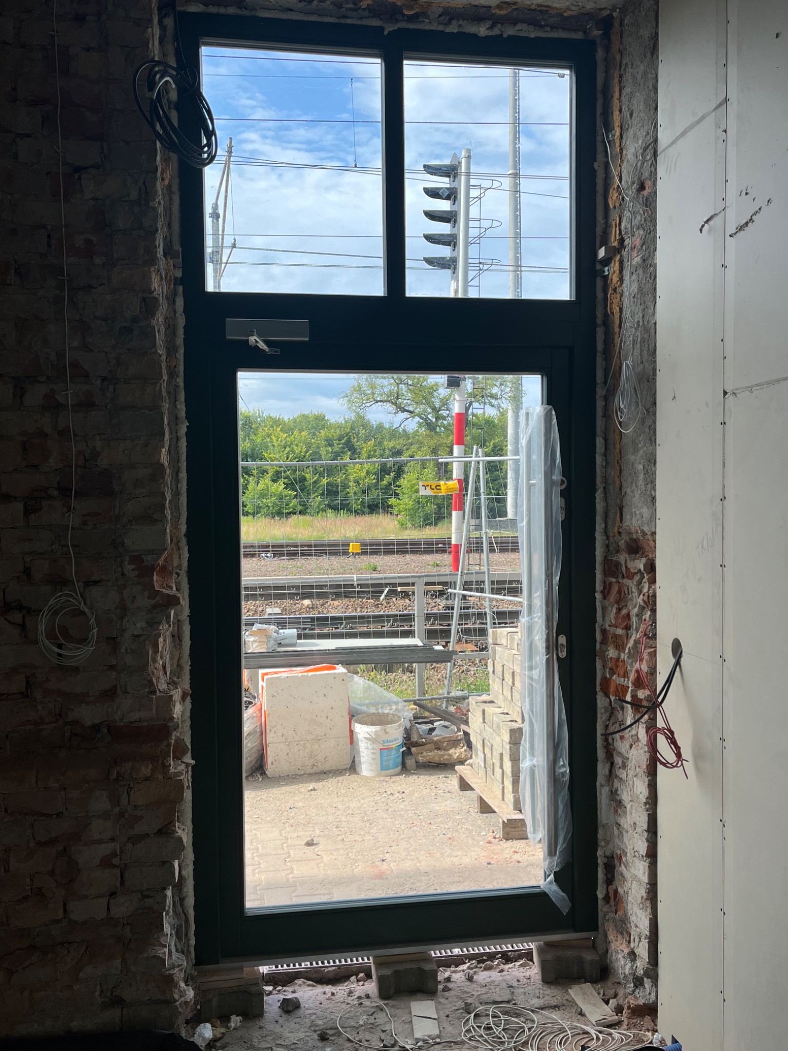 Surowy montaż okna z widokiem na tory kolejowe i sygnalizację, ściany w stanie deweloperskim, widoczne przewody i materiały budowlane.