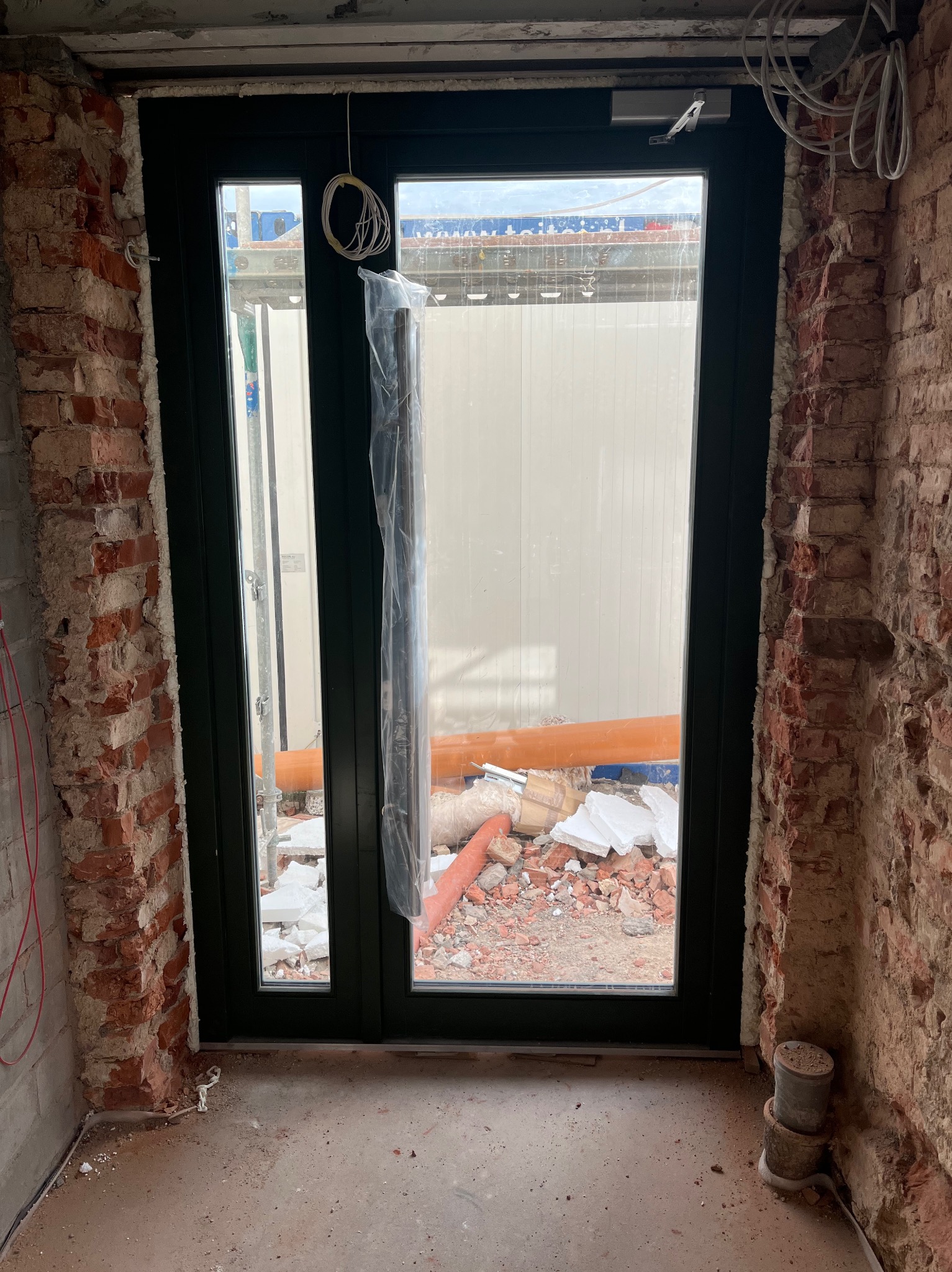 Montaż czarnych drzwi zewnętrznych w surowym otworze z cegły, widoczne przewody i elementy budowlane na zewnątrz.