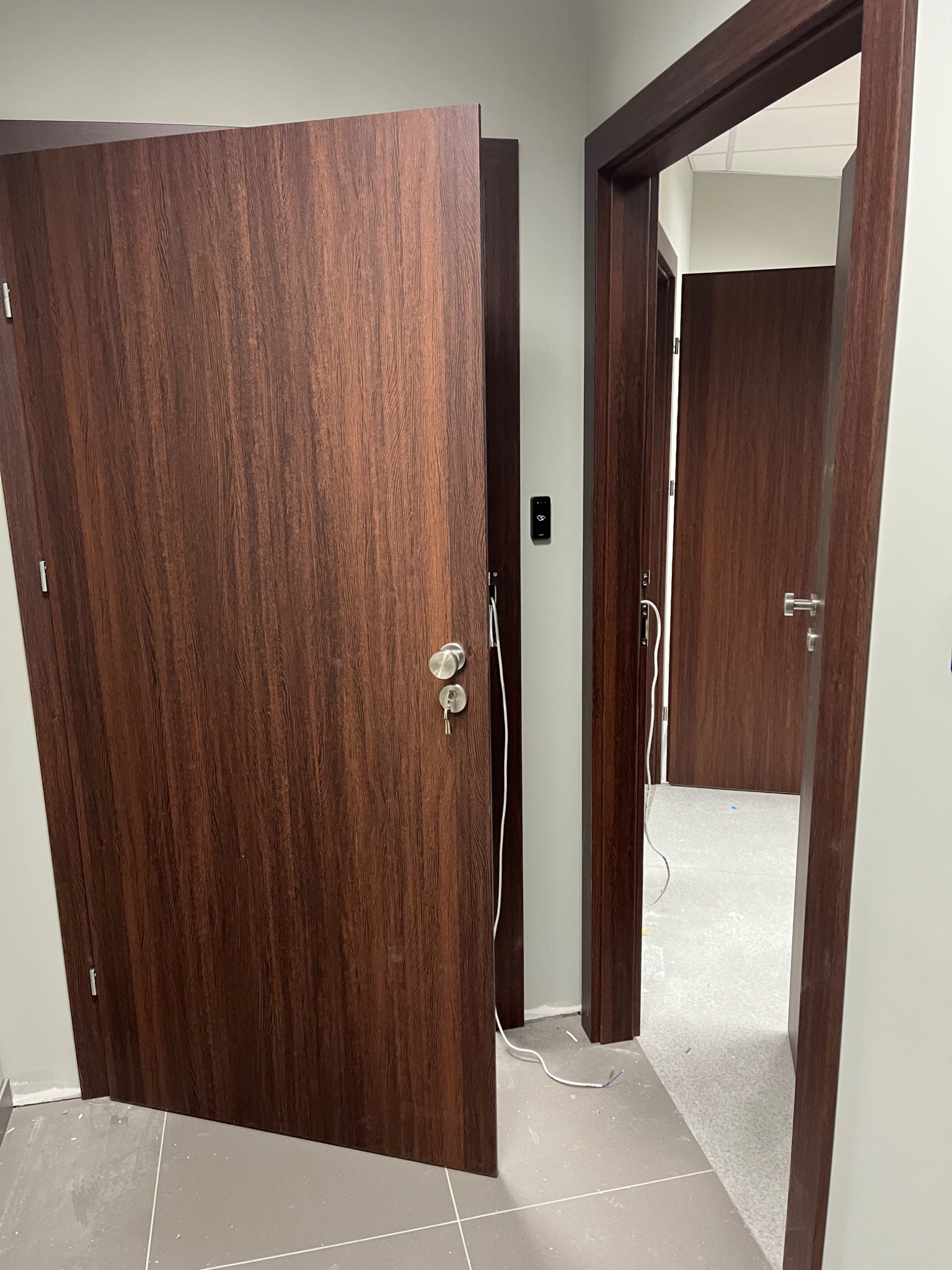 Brązowe drzwi wewnętrzne z widocznymi kablami podczas montażu, z kluczem w zamku, w tle widoczne kolejne drzwi i szara podłoga w trakcie wykończenia.
