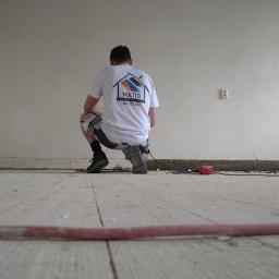 MATID-Mateusz Dworczak - Pracownik w białej koszulce z logo firmy MATID klęczy na drewnianej podłodze w trakcie prac remontowych, widoczny kabel i narzędzia, biała ściana w tle.