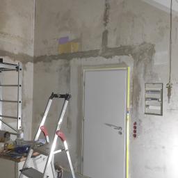 MATID-Mateusz Dworczak - Przygotowanie ścian do prac wykończeniowych: surowe ściany z widocznymi śladami po instalacjach, zabezpieczone taśmą malarską drzwi, rozstawiona drabina i widoczna tablica rozdzielcza elektryczna.