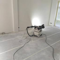 MATID-Mateusz Dworczak - Wnętrze w trakcie prac wykończeniowych: szpachlowane ściany i sufit z widocznymi łączeniami płyt, na podłodze taśmy maskujące i odkurzacz przemysłowy.