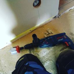 MATID-Mateusz Dworczak - Wiertarka Bosch z wiertłem koronowym leżąca na podłodze obok ściany z wywierconym otworem, widok z góry na etap prac wykończeniowych.