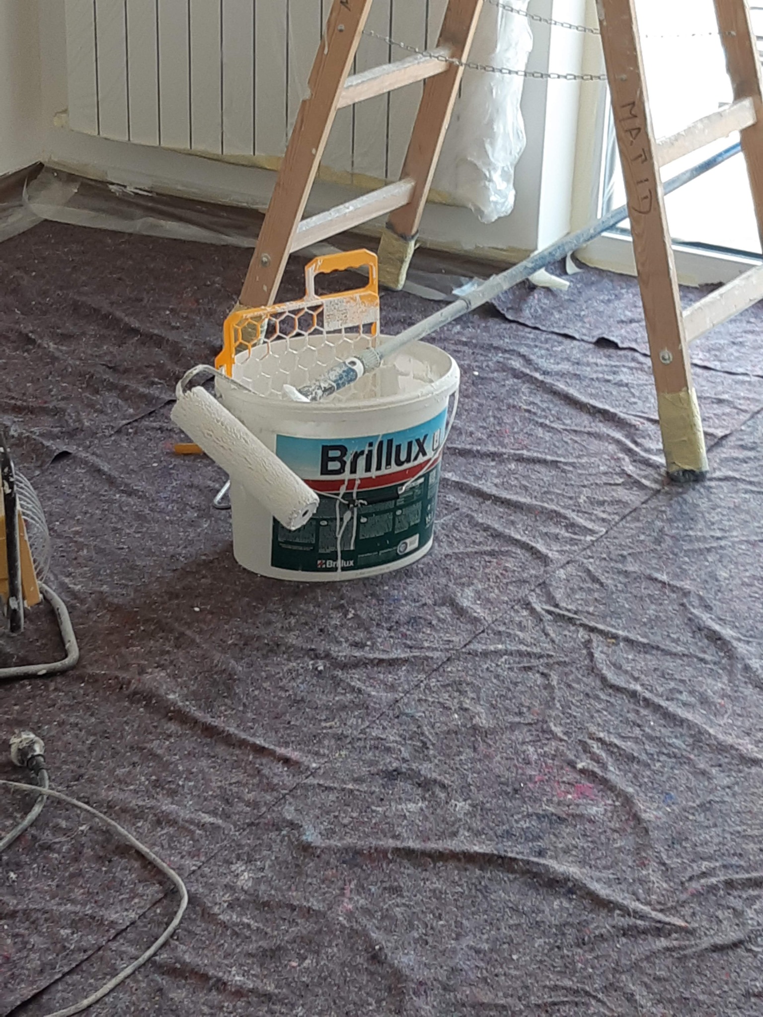 Przygotowania do malowania: wiadro z farbą Brilux, wałek do malowania, drabina malarska i folia ochronna na podłodze.