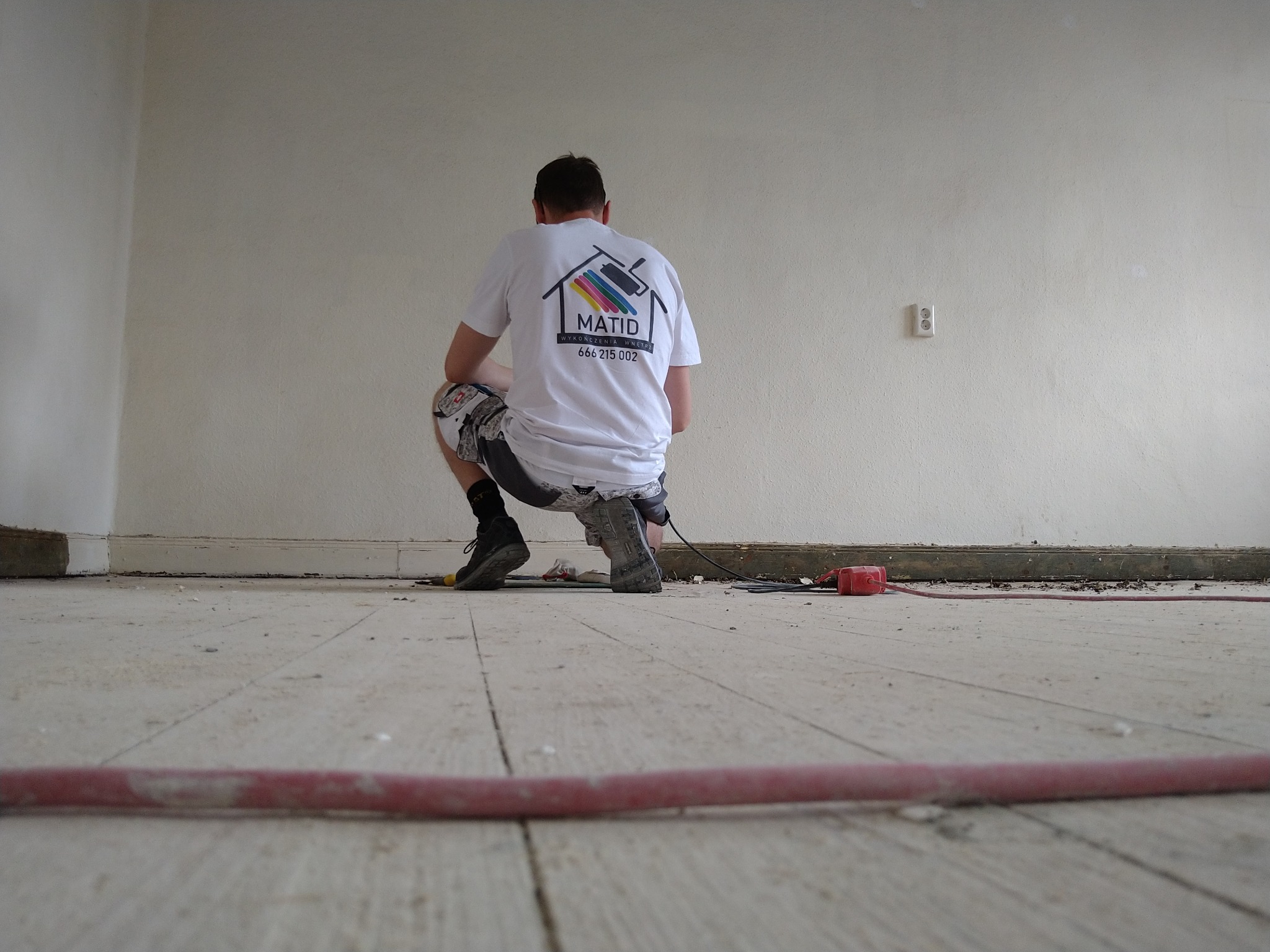 Pracownik w białej koszulce z logo firmy MATID klęczy na drewnianej podłodze w trakcie prac remontowych, widoczny kabel i narzędzia, biała ściana w tle.