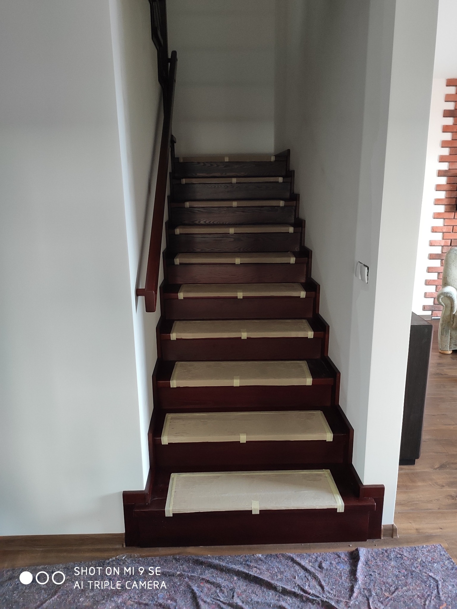 Schody drewniane w trakcie zabezpieczania folią malarską przed pracami wykończeniowymi, widok z dołu na górę, z ciemną balustradą i fragmentem ściany z cegły dekoracyjnej w tle.