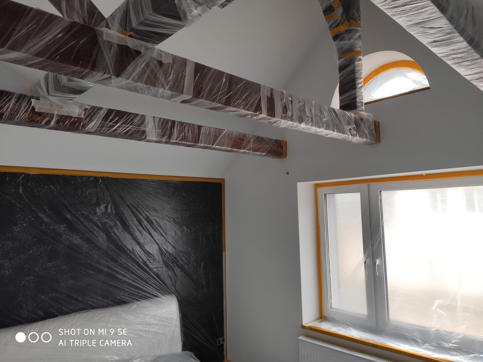 Pokój na poddaszu w trakcie zabezpieczania przed pracami remontowymi: drewniane belki stropowe owinięte folią, okna i framugi oklejone taśmą malarską, ściana zabezpieczona czarną folią malarską.