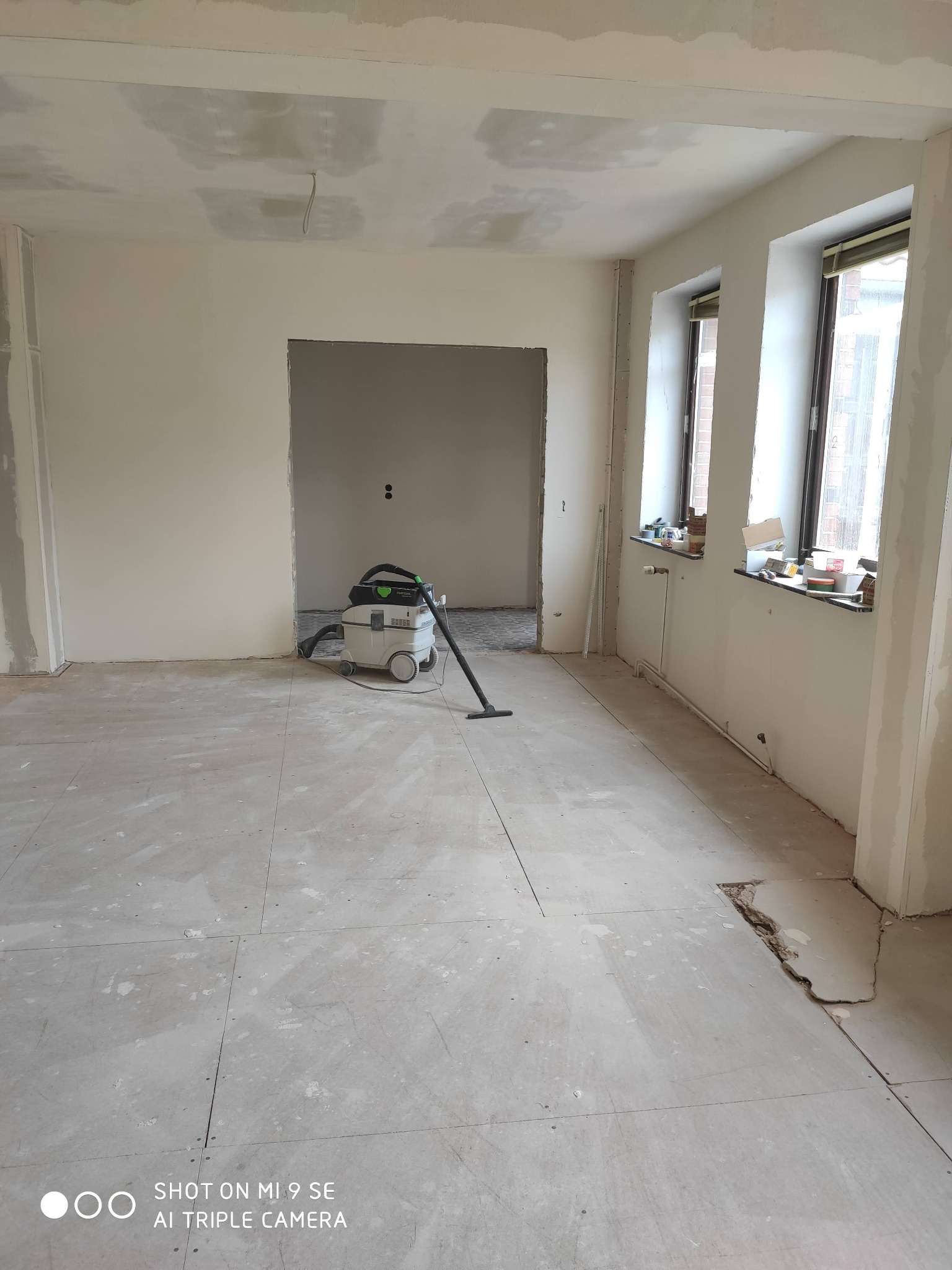 Pokój w trakcie remontu: widoczne płyty kartonowo-gipsowe na ścianach, podłoga przygotowana do położenia parkietu lub paneli, odkurzacz przemysłowy Festool, okna z parapetami zastawionymi...