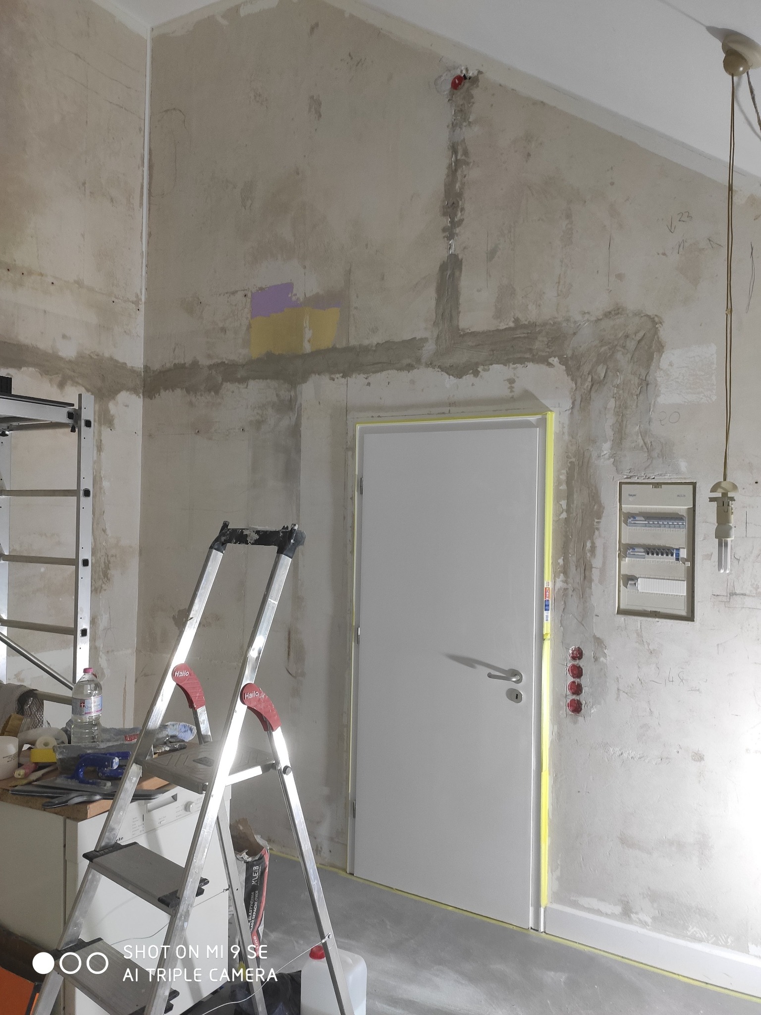 Przygotowanie ścian do prac wykończeniowych: surowe ściany z widocznymi śladami po instalacjach, zabezpieczone taśmą malarską drzwi, rozstawiona drabina i widoczna tablica rozdzielcza elektryczna.