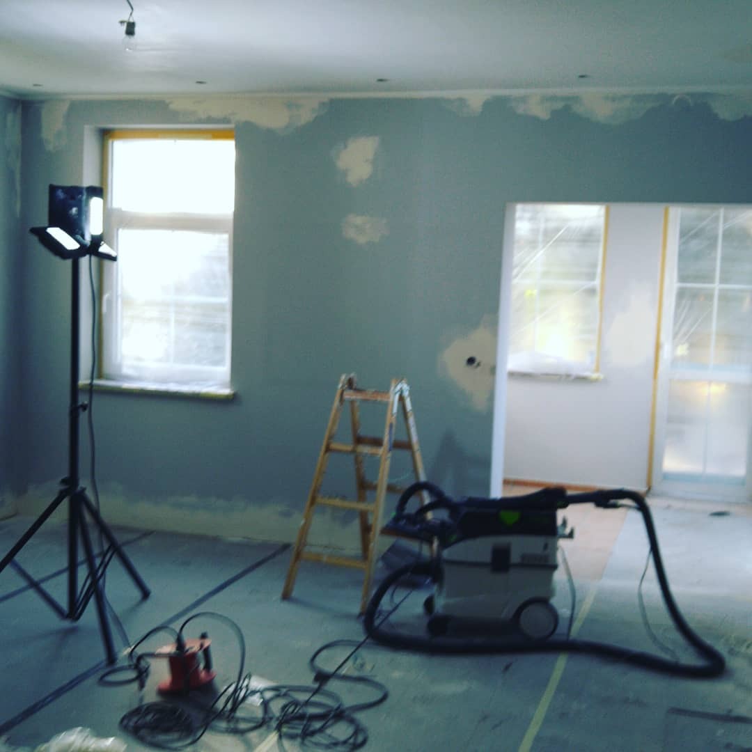 Pokój w trakcie remontu: szpachlowane ściany w odcieniach szarości, drabina, odkurzacz przemysłowy Festool i lampa budowlana na statywie.