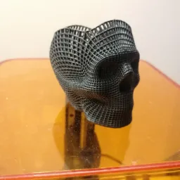 Czarna, ażurowa czaszka wydrukowana w technologii 3D, stojąca na pomarańczowej platformie drukarki 3D.