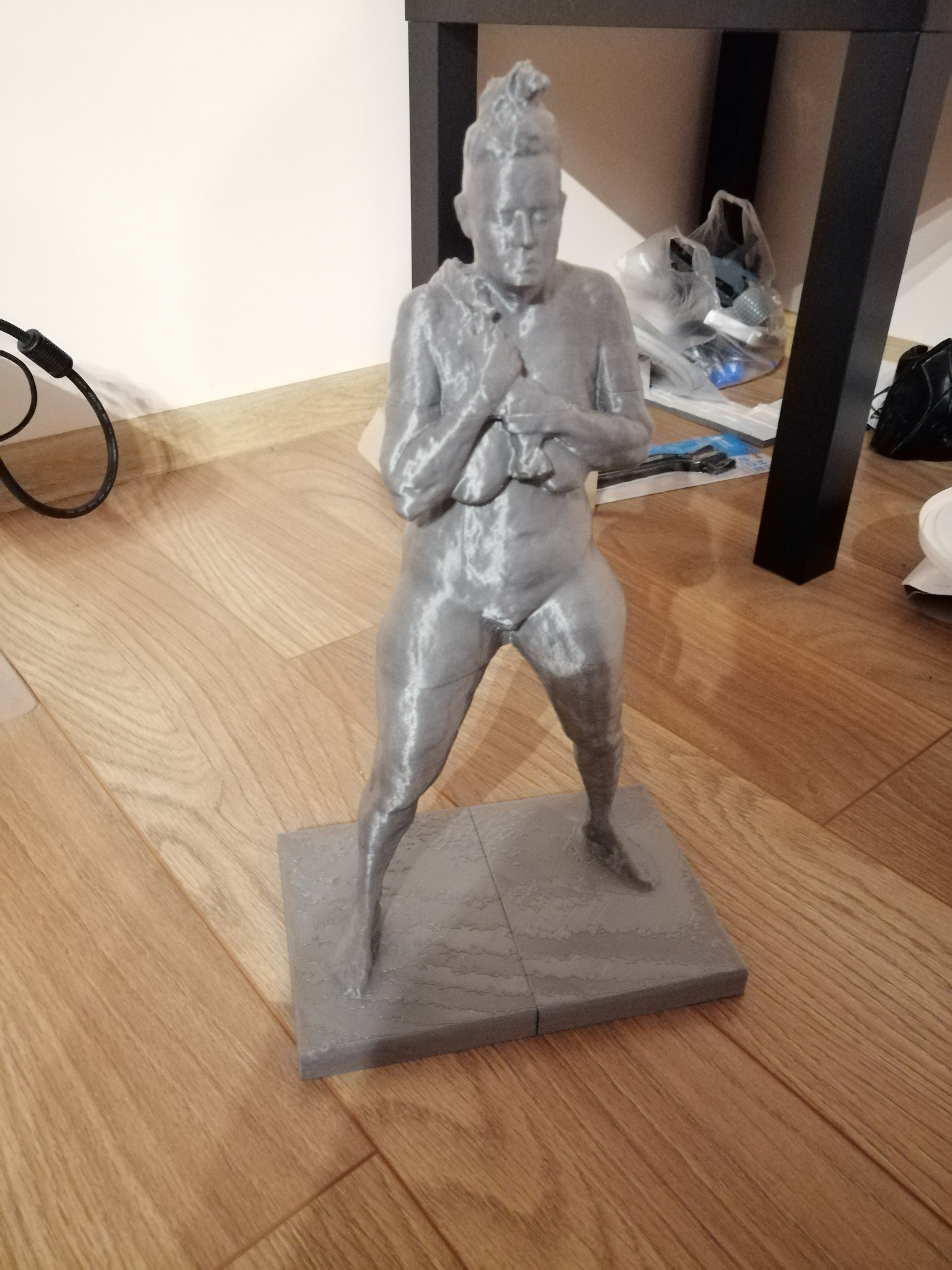Szara, wydrukowana w 3D figurka kobiety na podstawie, stojąca na drewnianej podłodze, z widocznymi śladami warstw druku.