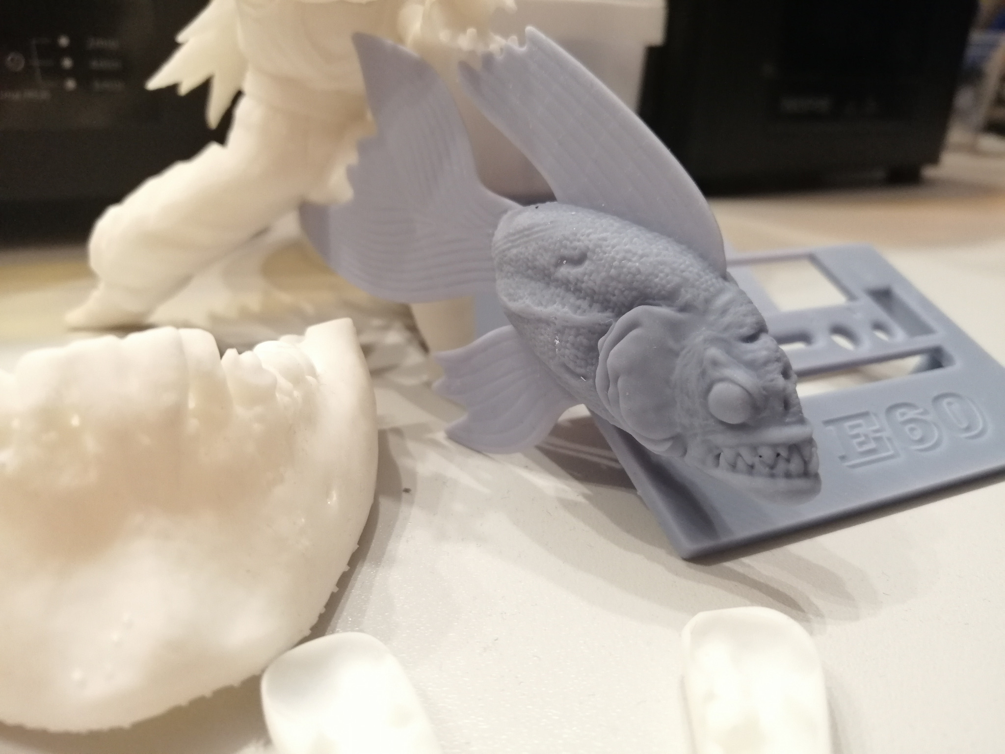 Niedokończone wydruki 3D w kolorze białym i szarym, przedstawiające m.in. zębatą rybę i fragmenty większej figury, leżące na stole obok platformy drukarki 3D z napisem E60.