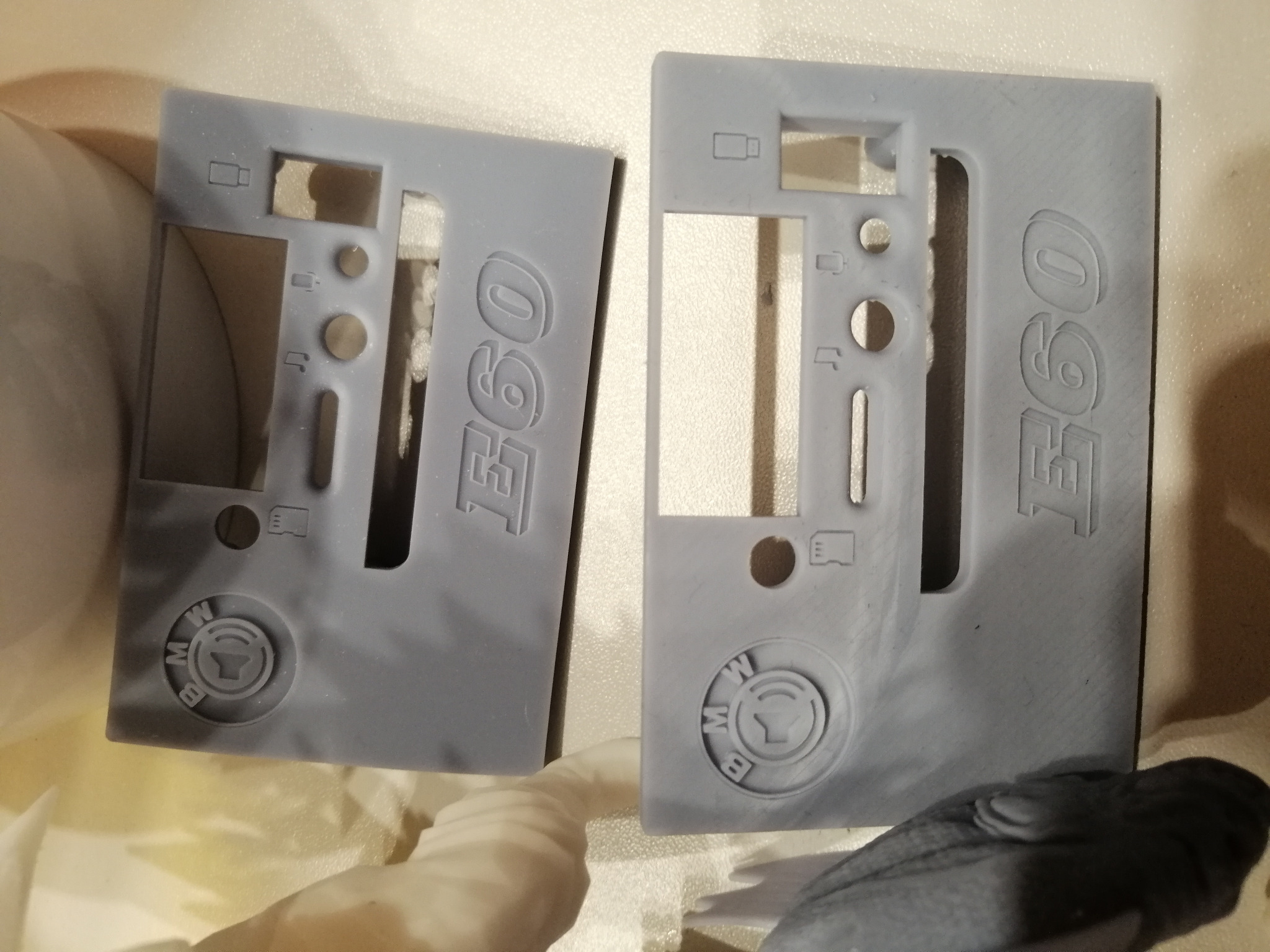 Dwa szare elementy z tworzywa sztucznego z wytłoczonym napisem 'E60' i logo BMW, prawdopodobnie prototypy lub części obudowy, leżące na jasnym tle.