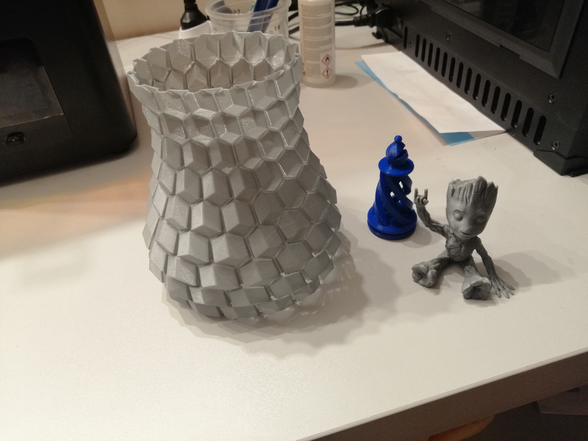 Trzy obiekty wydrukowane w 3D: wysoka, szara struktura o wzorze plastra miodu, niebieska, spiralna wieża i figurka Groota w szarym kolorze, wszystkie stoją na białym biurku obok drukarki 3D.