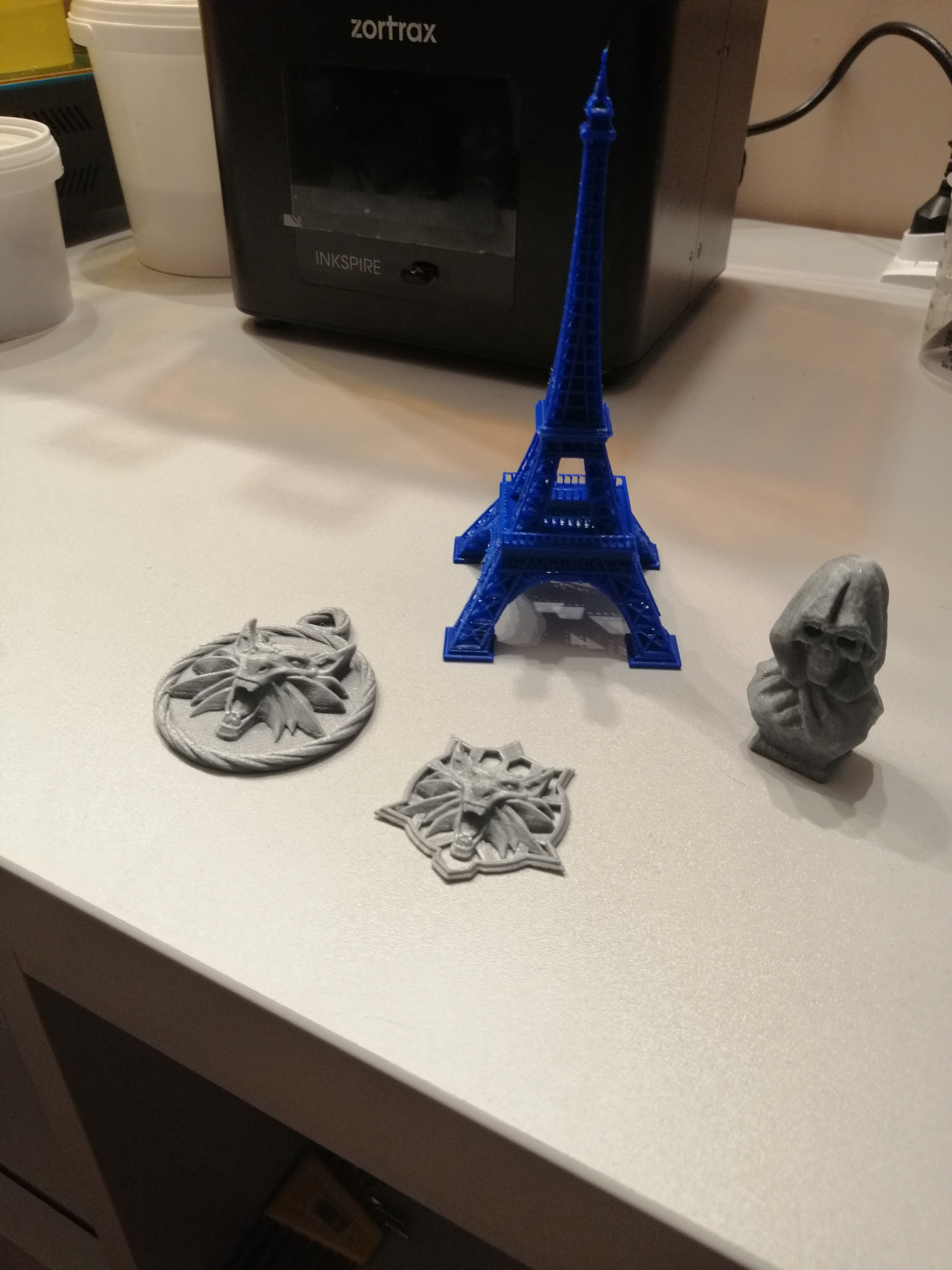 Niebieska miniatura wieży Eiffla oraz szare figurki, w tym medaliony z motywem wilka, wydrukowane na drukarce 3D Zortrax Inkspire.