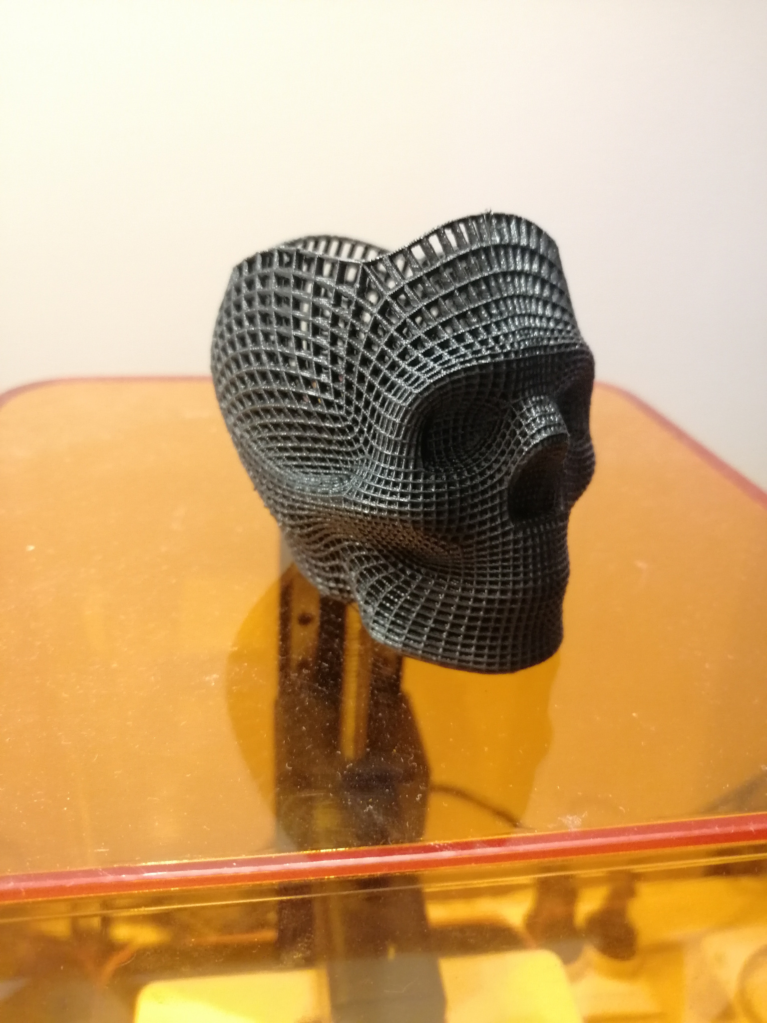 Czarna, ażurowa czaszka wydrukowana w technologii 3D, stojąca na pomarańczowej platformie drukarki 3D.