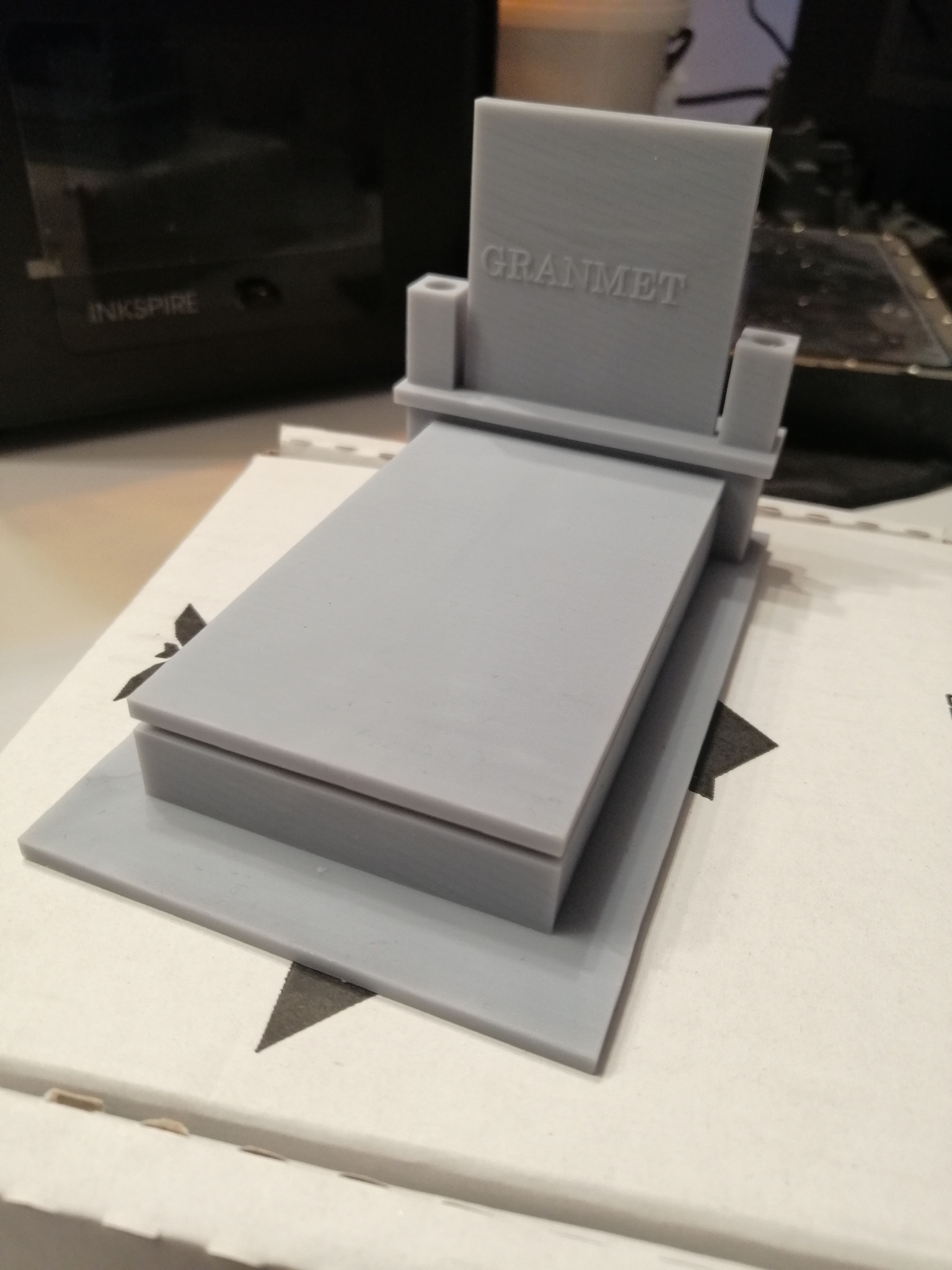 Szary, trójwymiarowy model nagrobka z napisem 'GRANMET', wydrukowany na drukarce 3D, widoczna drukarka Inkspire w tle.