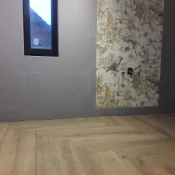 Ściana wyłożona szarymi płytkami z wąskim oknem, obok fragment ściany z tapetą w kwiaty i ptaki, na podłodze parkiet ułożony w jodełkę.