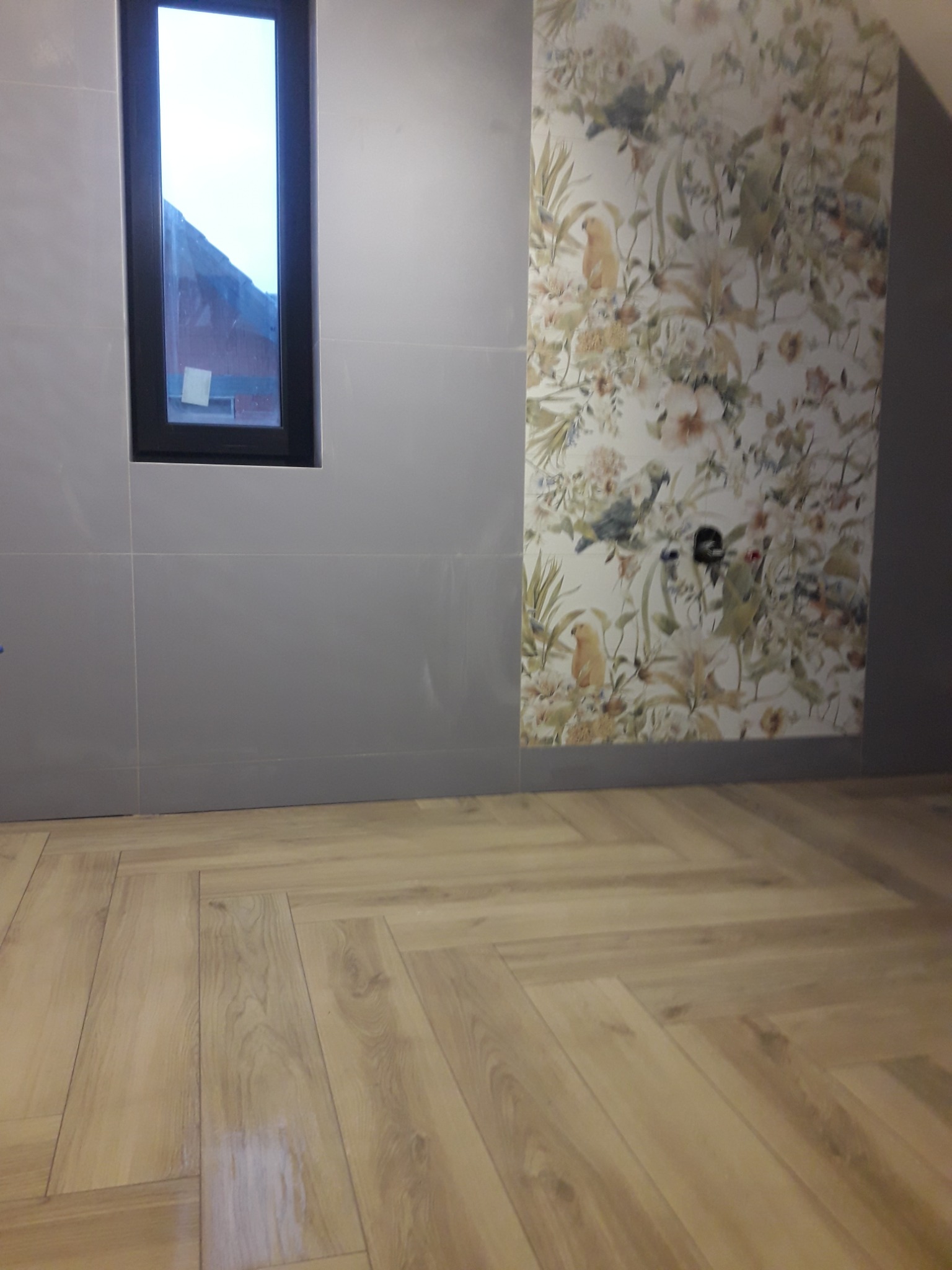 Ściana wyłożona szarymi płytkami z wąskim oknem, obok fragment ściany z tapetą w kwiaty i ptaki, na podłodze parkiet ułożony w jodełkę.
